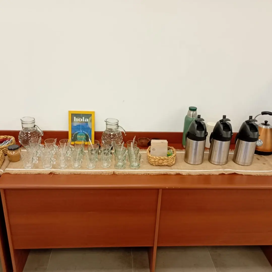 Termos de acero inoxidable para café, pava eléctrica de cobre y tazas descartables sobre un mantel de arpillera en un break de oficina.