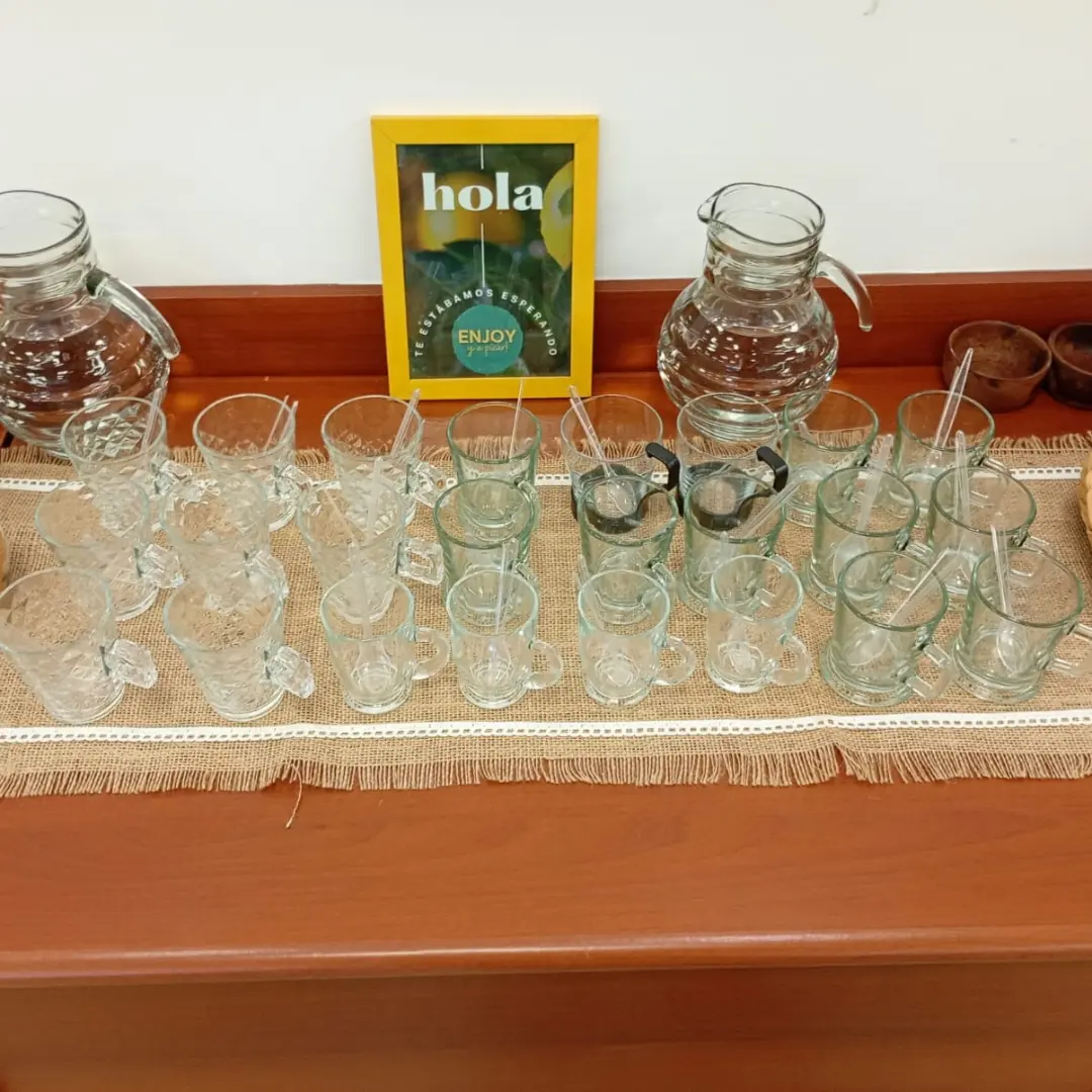 Estación de vajilla con tazas de vidrio para buffet corporativo junto a un cuadro amarillo con el logo de Enjoy y a picar.