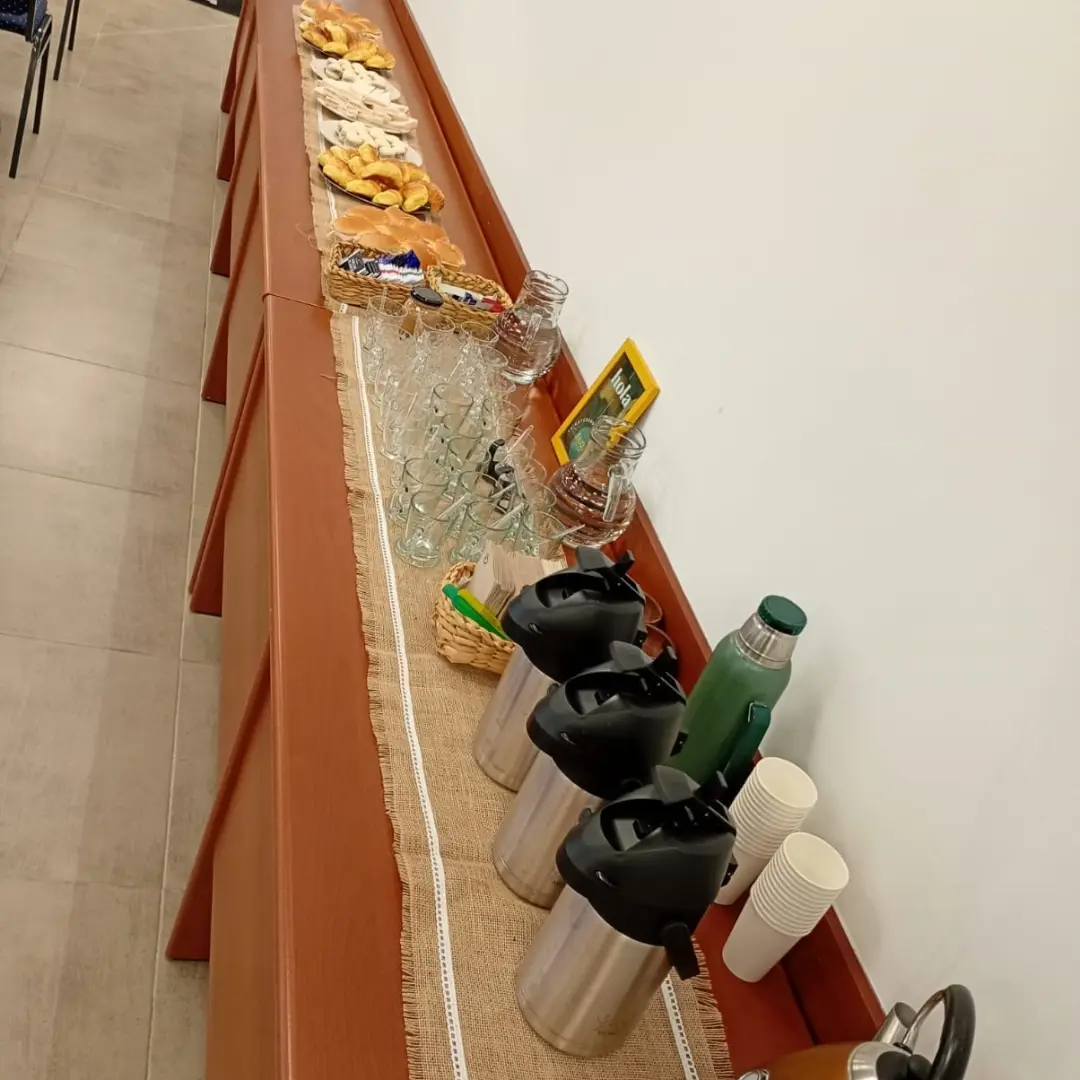 Termos de acero inoxidable para café, pava eléctrica de cobre y tazas descartables sobre un mantel de arpillera en un break de oficina.